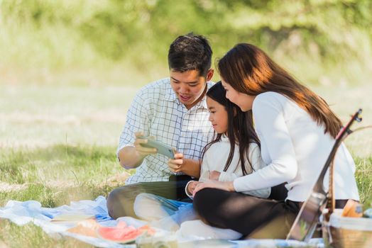 Happy Asian young family father, mother and child little girl having fun and enjoying outdoor sitting on picnic blanket looging picture after taking selfie by mobile smart phone at summer garden park