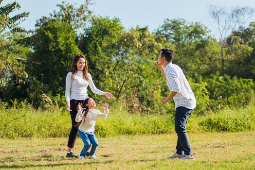 Happy Asian lifestyle family mother, father and little cute girl child having fun together and enjoying outdoor play blowing soap bubbles in the garden park on a sunny day, summertime