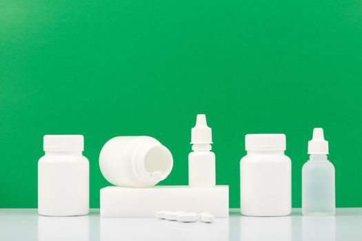 White plastic bottles with medications, pills, nose spray, ear drops and eye drops on white table against green background with copy space. Concept of health care, medical treatment and pharmacy