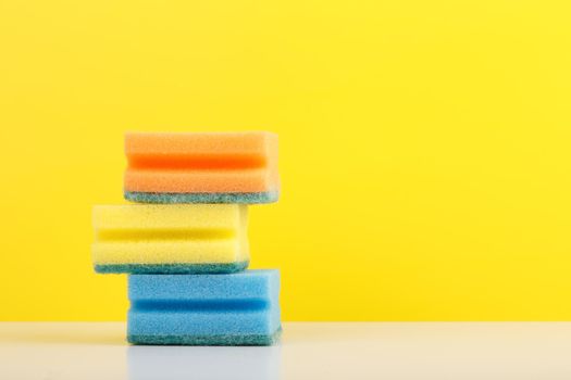 Blue, yellow and orange sponges for washing dishes on white table against yellow background with copy space. Concept of house cleaning