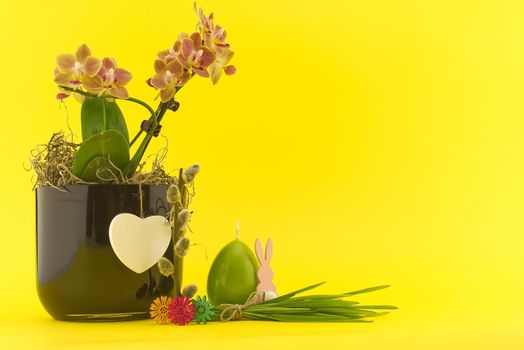 Easter Decoration with flowers, egg shaped candle, pussy willow branch and bundle of wheat seedlings over a yellow background. Happy Easter card concept with free copy space for text