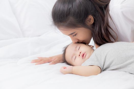 Young asian mother kiss cheek of little baby girl with tender on bed in the bedroom, mom love newborn and care, mother with expression with child together, parent and daughter, family concept.