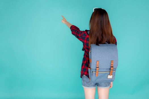 Back view of beautiful young asian woman travel with backpack pointing something isolated on blue background, rear asia girl is student or tourist in summer trip, holiday concept.