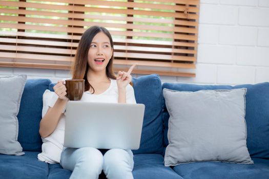 Young asian business woman smile and work from home with laptop computer online on sofa in living room, freelance girl using notebook and drink coffee on couch, new normal, lifestyle concept.