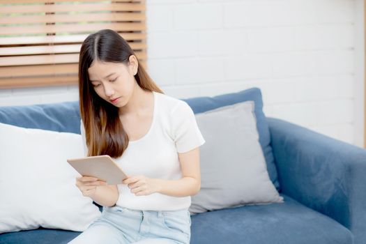 Young attractive asian woman resting using browsing tablet computer on sofa at home, happy girl sitting on couch relax reading digital gadget at house, communication and lifestyle concept.