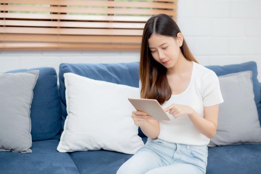 Young attractive asian woman resting using browsing tablet computer on sofa at home, happy girl sitting on couch relax reading digital gadget at house, communication and lifestyle concept.
