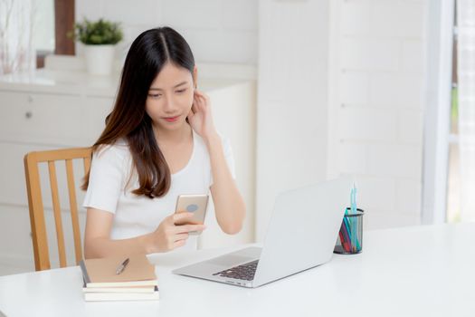 Young asian woman working laptop computer and reading smartphone on internet online on desk at home, freelance girl smiling using phone with social media, business and communication concept.