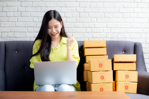 Young asian woman is merchant using laptop computer on sofa checking order of customer with purchase online shopping, business SME or startup, entrepreneur or seller and service delivery product.