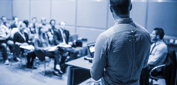 Speaker giving a talk on corporate business conference. Unrecognizable people in audience at conference hall. Business and Entrepreneurship event. Blue toned black and white.