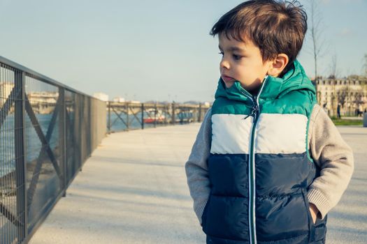 Portrait of little boy wearing sleeveless jacket, child looking away from camera, stands on promenande along river.