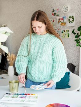 Young woman artist in mint green sweater holding color palette working at home