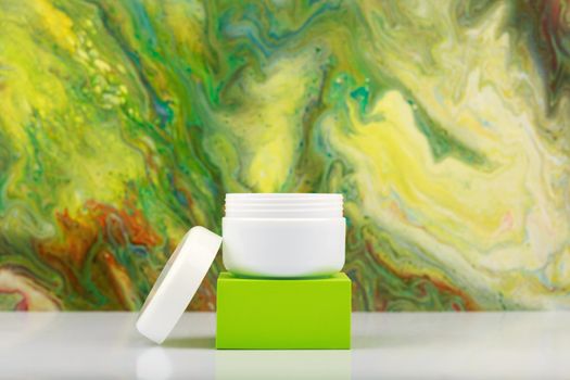 Opened cream jar on green pedestal against abstract background in bright green colors with copy space. Face cream, scrub or mask against colorful background.