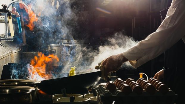 Chef stir fry the food in a frying pan, smoke and splatter the sauce in the kitchen.