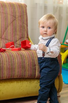 Little boy, child stands by the couch with a festive gift box. In a nice outfit, a white shirt and trousers with suspenders.
