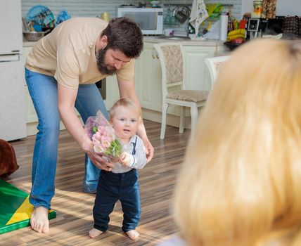 Dad with baby, son congratulate mom on the holiday. The kid brings his mother a bouquet of flowers and a gift.