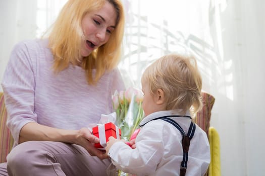 The kid congratulates his mother on the holiday, gives her flowers and a gift. Boy in white shirt and trousers with suspenders