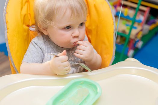 Little child sits on a high chair and eats with a spoon. Close-up