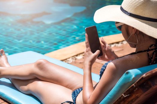 Young beautiful girl woman in bikini sitting on chair near pool blue water on resort using in mobile phone