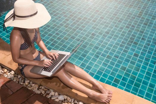 Asian Young woman in big hat relaxing on poolside with laptop computer blue water