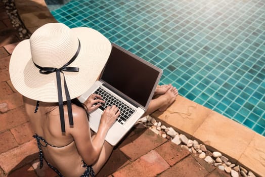 Asian Young Woman using her laptop with big hat on poolside edge on sunny day blue water