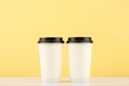Two white tall disposable coffee or tea cups on white table against light yellow background with copy space. Hot drinks minimalistic concept