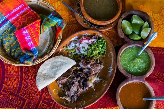 Top view of chopped lamb meat, hot sauces, lime slices, broth, and tortillas on colorful tablecloth. Tasty meal and condiments in clay plates and baskets above red and orange table. Mexican cuisine