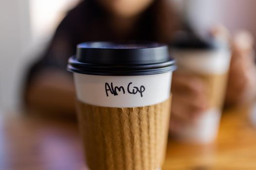 Close-up of white and brown paper coffee cup with the blurry background. Disposable cup on wooden table with black plastic cap and the flavor written on it. Hot food and drinks
