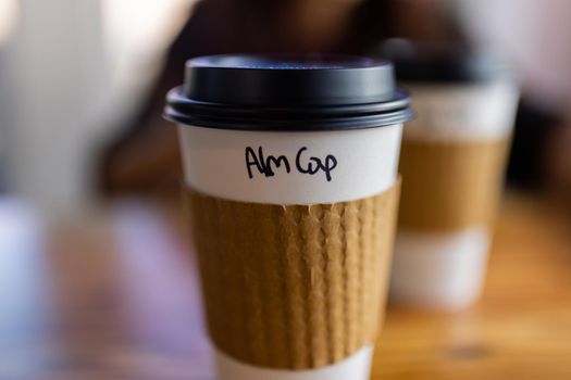Close-up of white and brown paper coffee cup with the blurry background. Disposable cup on wooden table with black plastic cap and the flavor written on it. Hot food and drinks