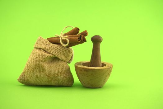 Cinnamon sticks bundle tied with jute string on top of hessian bag and wooden mortar with pestle over green background and free copy space for text