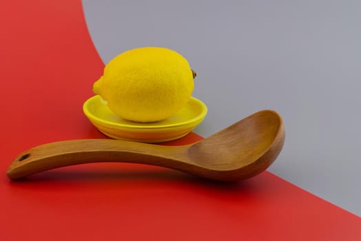 Lemon on a yellow plate next to a wooden spoon on a two tone grey and red background with copy space. Color trends concept
