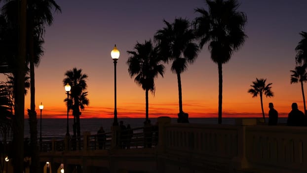 Oceanside, California USA - 27 Dec 2020: Palms silhouette twilight sky, dusk nightfall atmosphere. People walking on pier. Tropical pacific ocean beach, sunset afterglow aesthetic. Los Angeles vibes.