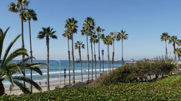 Oceanside, California USA - 8 Feb 2020: Pacific ocean beach, green palm trees and pier. Sunny day, tropical waterfront resort. Vista point near Los Angeles. Summer sea coast aesthetic, people walking.