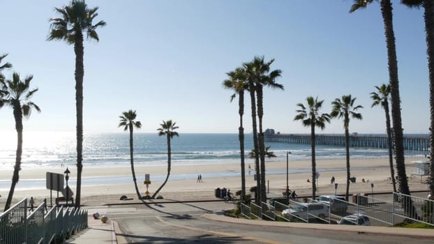 Oceanside, California USA - 8 Feb 2020: People walking strolling, waterfront resort street, pacific ocean tropical beach with palm trees. Person riding bicycle, coastal road perspective. Biker cycling