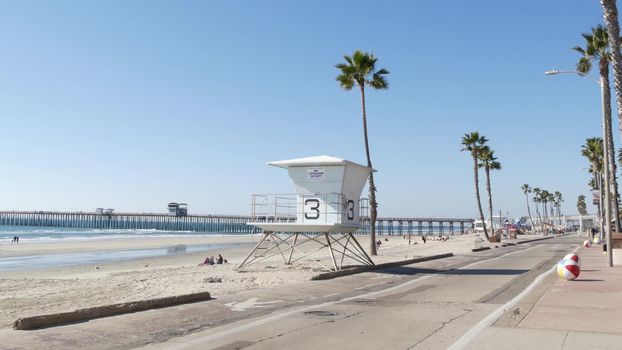 Oceanside, California USA - 8 Feb 2020: People walking, waterfront promenade near pier, pacific ocean tropical beach tourist resort with palm trees. Lifeguard tower. Disabled senior man in wheelchair.