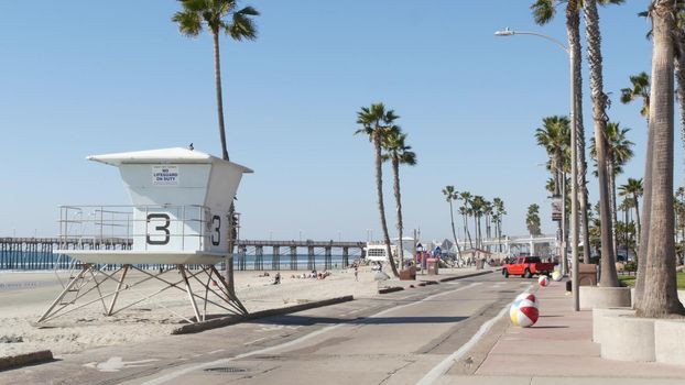 Oceanside, California USA - 8 Feb 2020: People walking, waterfront promenade near pier, pacific ocean tropical beach tourist resort with palm trees. Lifeguard tower on beachfront street on sunny day.