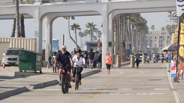 Oceanside, California USA - 8 Feb 2020: People walking, waterfront promenade, beachfront boardwalk near pier. Vacations ocean beach resort near Los Angeles. Biker riding bicycle, senior couple cycling