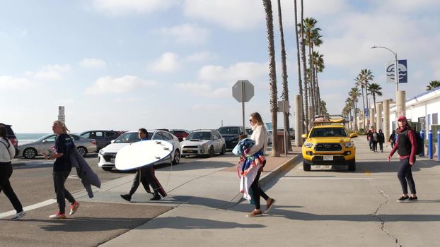 Oceanside, California USA - 8 Feb 2020: Women walking on waterfront promenade, people on beachfront boardwalk. Vacations ocean beach resort near Los Angeles. Surfers with surfboards, lifeguard car.