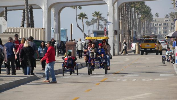 Oceanside, California USA - 8 Feb 2020: People walking on waterfront promenade, beachfront boardwalk. Vacations ocean beach resort near Los Angeles. Family riding surrey 4 wheel double bench bike.