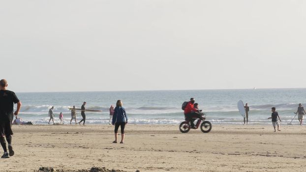 Oceanside, California USA - 8 Feb 2020: People walking on ocean beach, waterfront vacations resort. Surfer men with surfboard going surfing in sea waves. Family riding bikes. Man with metal detector.