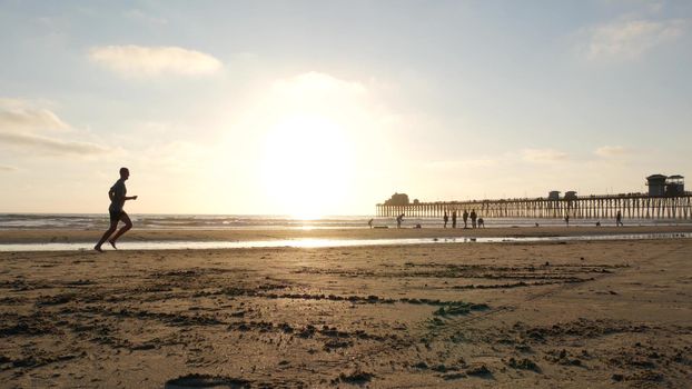 Oceanside, California USA - 8 Feb 2020: People walking on ocean beach, waterfront vacations resort. Contrast silhouette, man running, sand near sea waves. Young guy training in sunset light near pier.