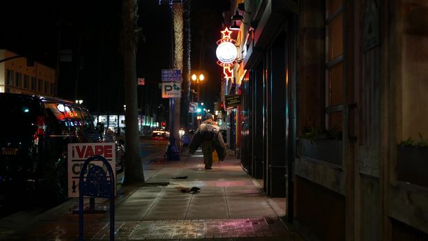 Oceanside, California USA - 9 Feb 2020: Lonely man looks like homeless walking, city street walkway at night. Male person on sidewalk, possible unemployed jobless beggar. Poverty and begging problem.