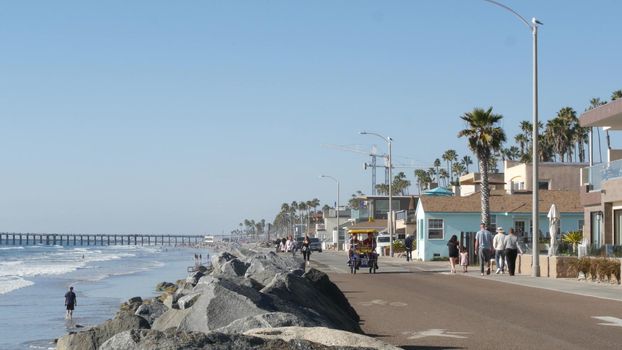 Oceanside, California USA - 16 Feb 2020: People walking strolling on waterfront sea promenade, beachfront boardwalk near pier. Vacations ocean beach resort near Los Angeles. Double bench surrey bike.