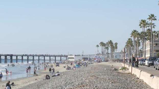 Oceanside, California USA -16 Feb 2020: Many people walking strolling on waterfront sea promenade, beachfront near pier. Vacations ocean beach tourist resort near Los Angeles. Crowd by lifeguard tower