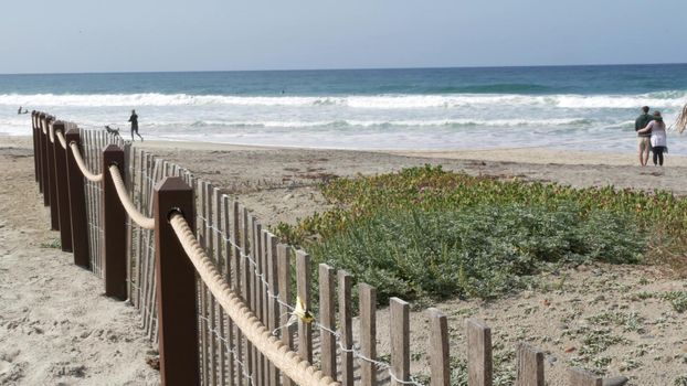 Encinitas, California USA - 23 Feb 2020: Pacific coast, people walking on ocean beach by sea water waves. Coastal access with picket fence on sandy shore near San Diego and Los Angeles. Couple and dog