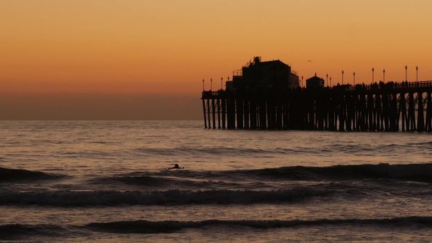 Oceanside, California USA - 16 Feb 2020: Surfer silhouette, pacific ocean beach in evening, water waves and sunset. Tropical coastline, waterfront vacation resort. People enjoy surfing as sport hobby.