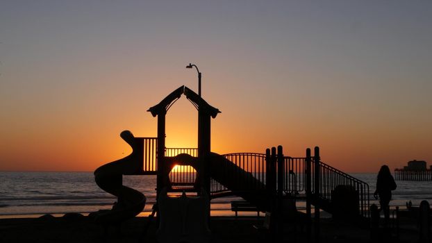 Oceanside, California USA - 11 Feb 2020: Silhouette of waterfront playground near pier. Children playing with parents, sunset ocean beach. Family with kids, beachfront recreation area, pacific coast.
