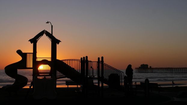 Oceanside, California USA - 11 Feb 2020: Silhouette of waterfront playground near pier. Children playing with parents, sunset ocean beach. Family with kids, beachfront recreation area, pacific coast.