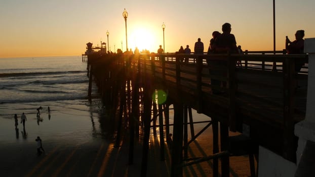 Oceanside, California USA - 17 Nov. 2019: Wooden pier and people walking. Tourists strolling in waterfront resort, summertime vacations near Los Angeles. Sunset ocean beach, tropical sunny orange sky.