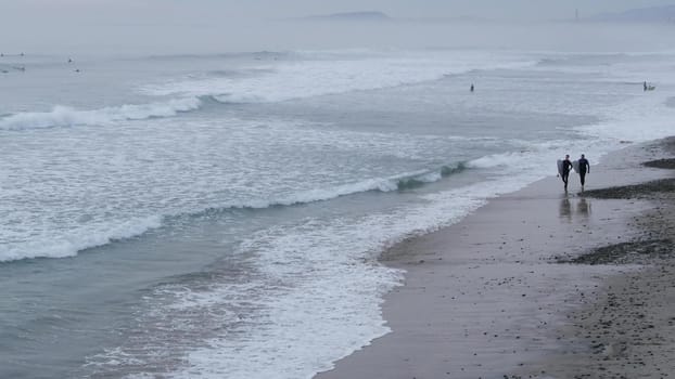 Encinitas, California USA - 21 Dec. 2019: People walking on beach after surfing, cold ocean water. Surfers near sea waves with surfboard. Aquatic sport hobby, misty winter weather. Healthy lifestyle.