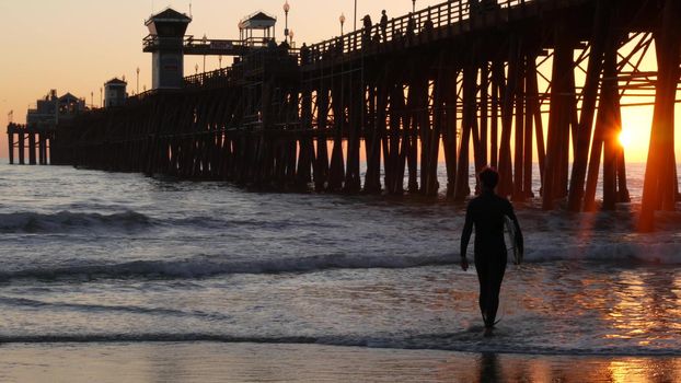 Oceanside, California USA - 16 Feb 2020: Surfer silhouette, pacific ocean beach in evening, water waves and sunset. Tropical coastline, waterfront vacation resort. People enjoy surfing as sport hobby.